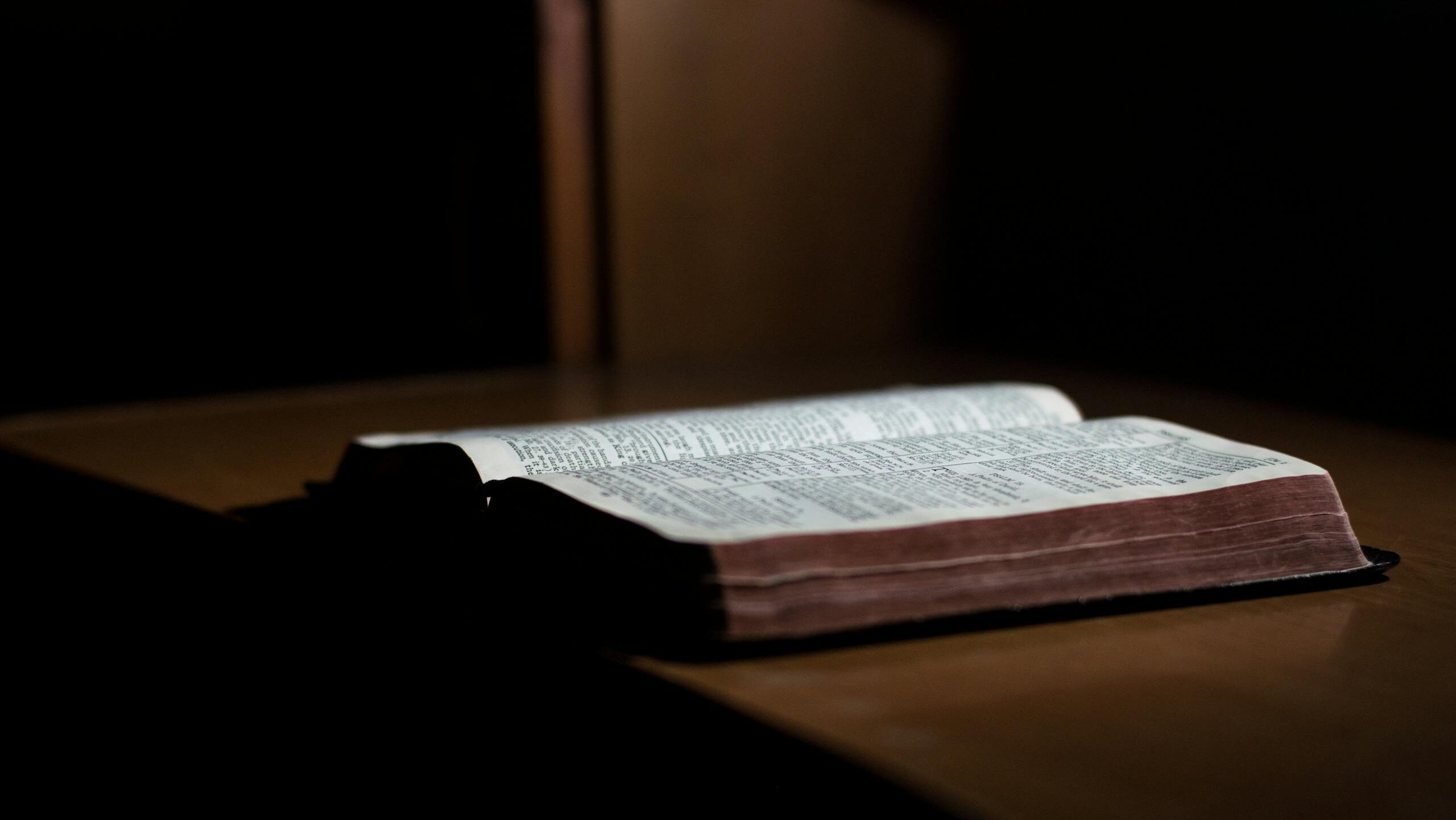 A dimly lit open Bible rests on a wooden table, inviting reflection and study.