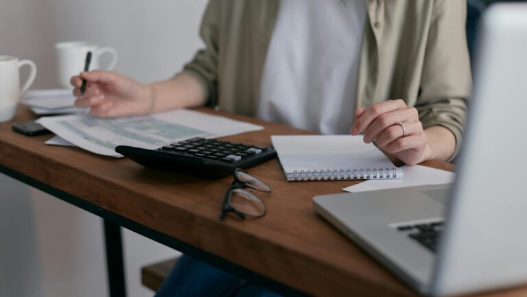 A woman manages finances at home, using a laptop and calculator on a wooden desk.