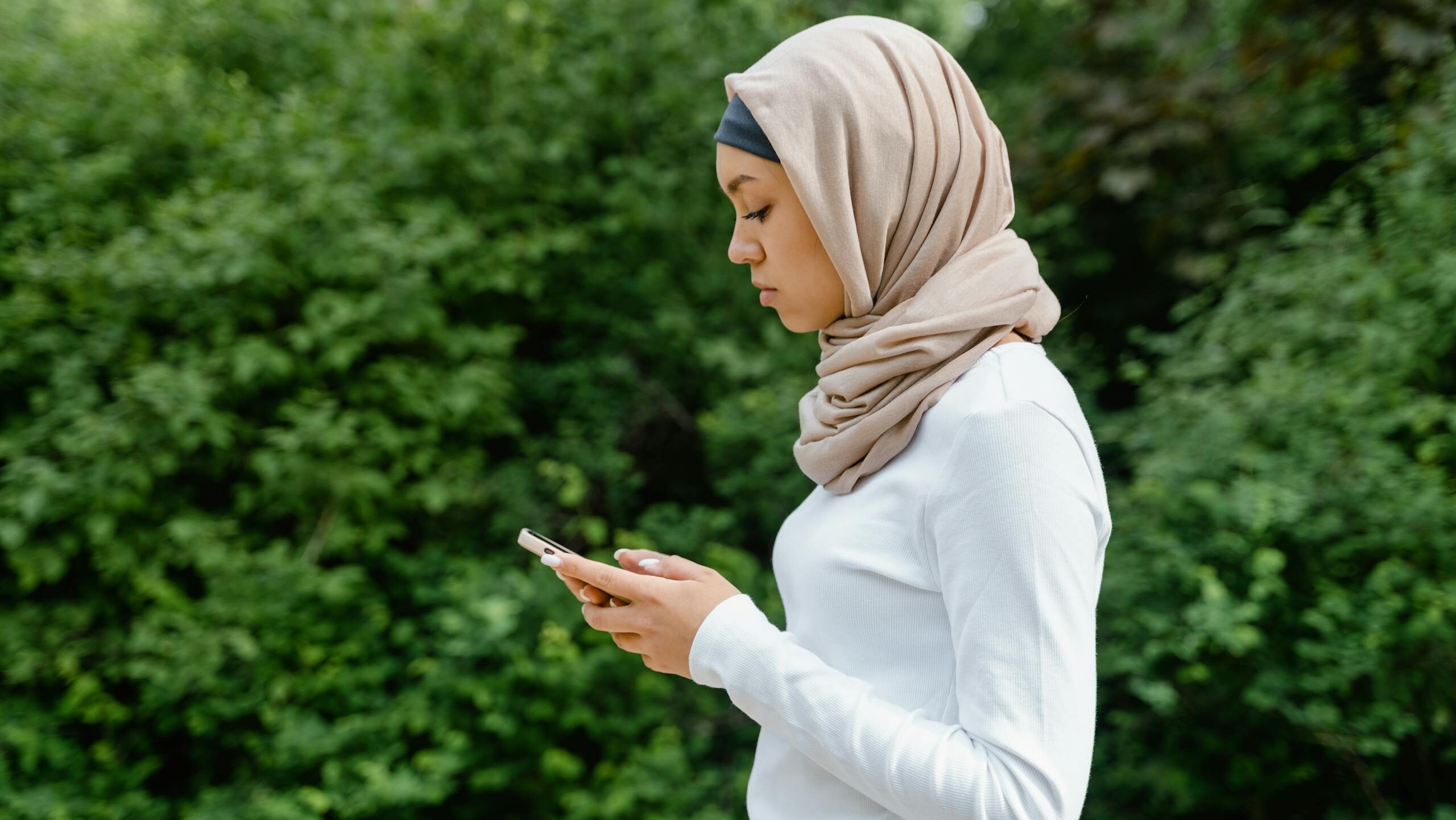Side view of a Muslim woman in hijab using a smartphone in a natural setting.
